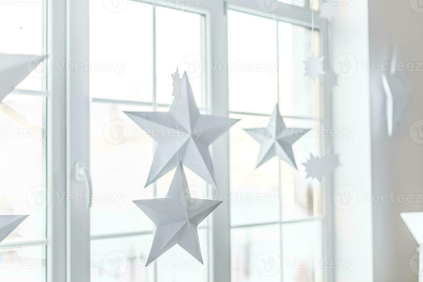 Large bright window. On the window-sill is a gray knitted plaid with gifts and sparkles with herlands. Paper Christmas stars are suspended on the background of the window. photo