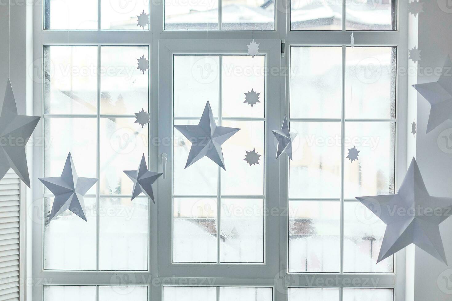 Large bright window. On the window-sill is a gray knitted plaid with gifts and sparkles with herlands. Paper Christmas stars are suspended on the background of the window. photo