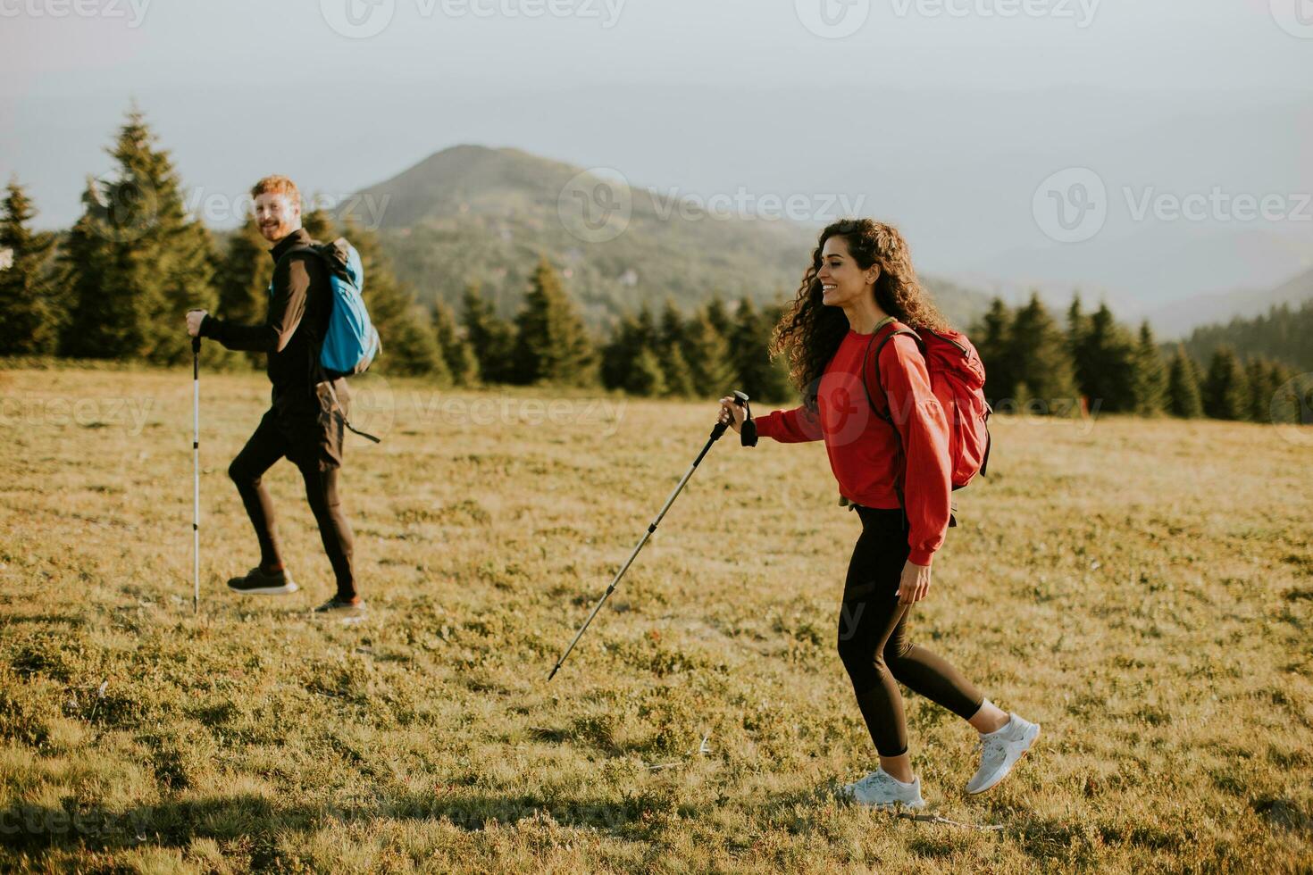 Smiling couple walking with backpacks over green hills photo
