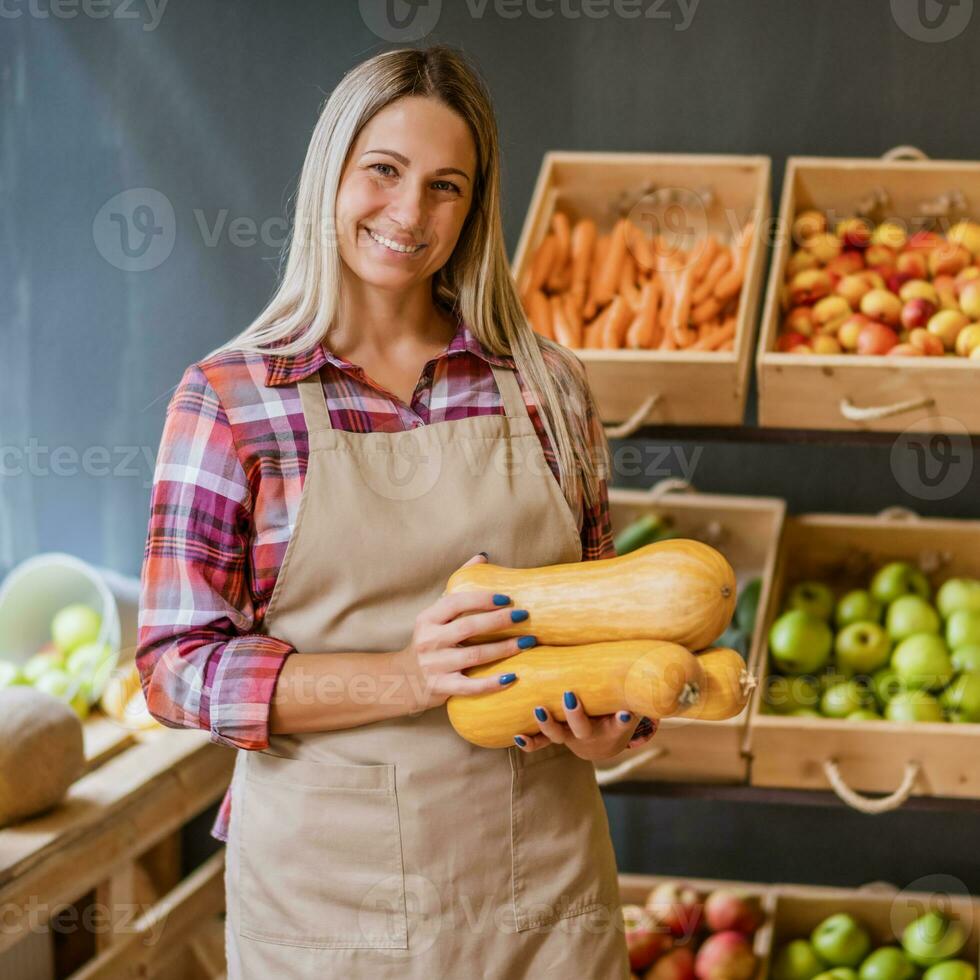 Woman works in fruits and vegetables shop. She is holding basket with butternut squash. photo