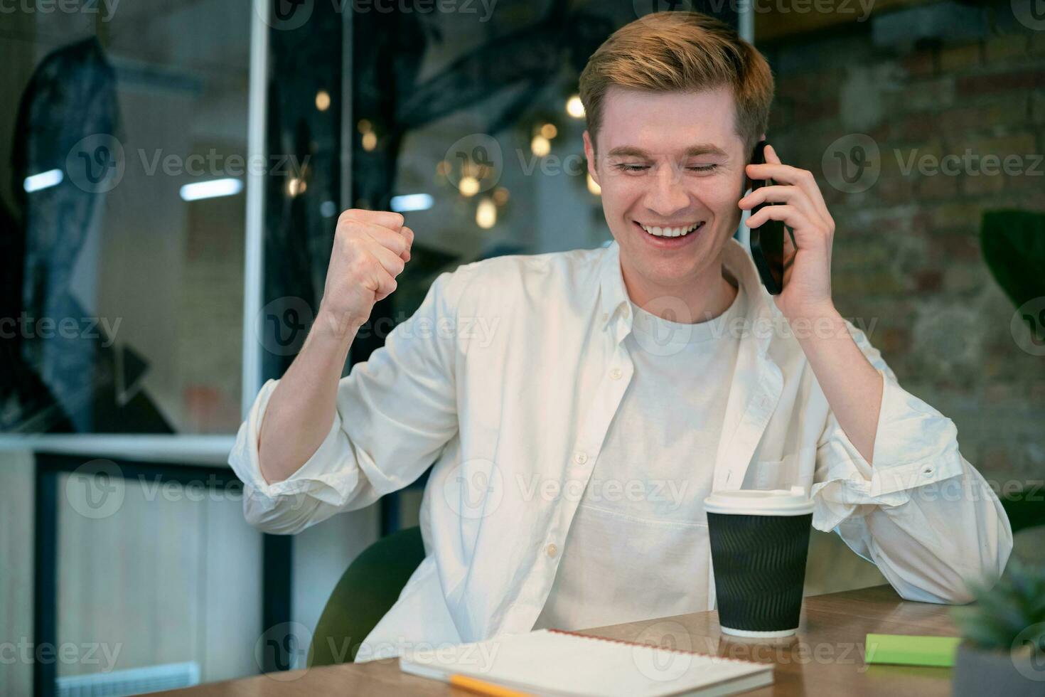 Happy young man holding mobile phone while talking, sitting at his workplace in modern office. Business, coworking, management concept photo