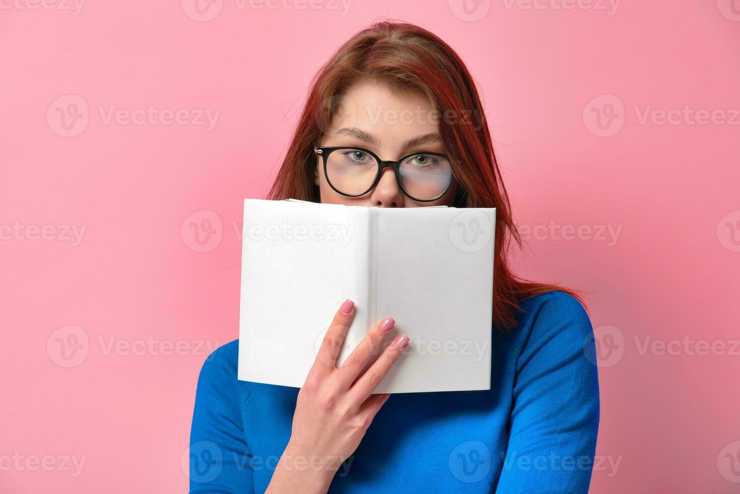 Portrait of a young charming girl with beautiful green eyes in glasses holding a book with a blank white cover, hiding lips while looking at camera. Isolated on pink colored background in studio. photo