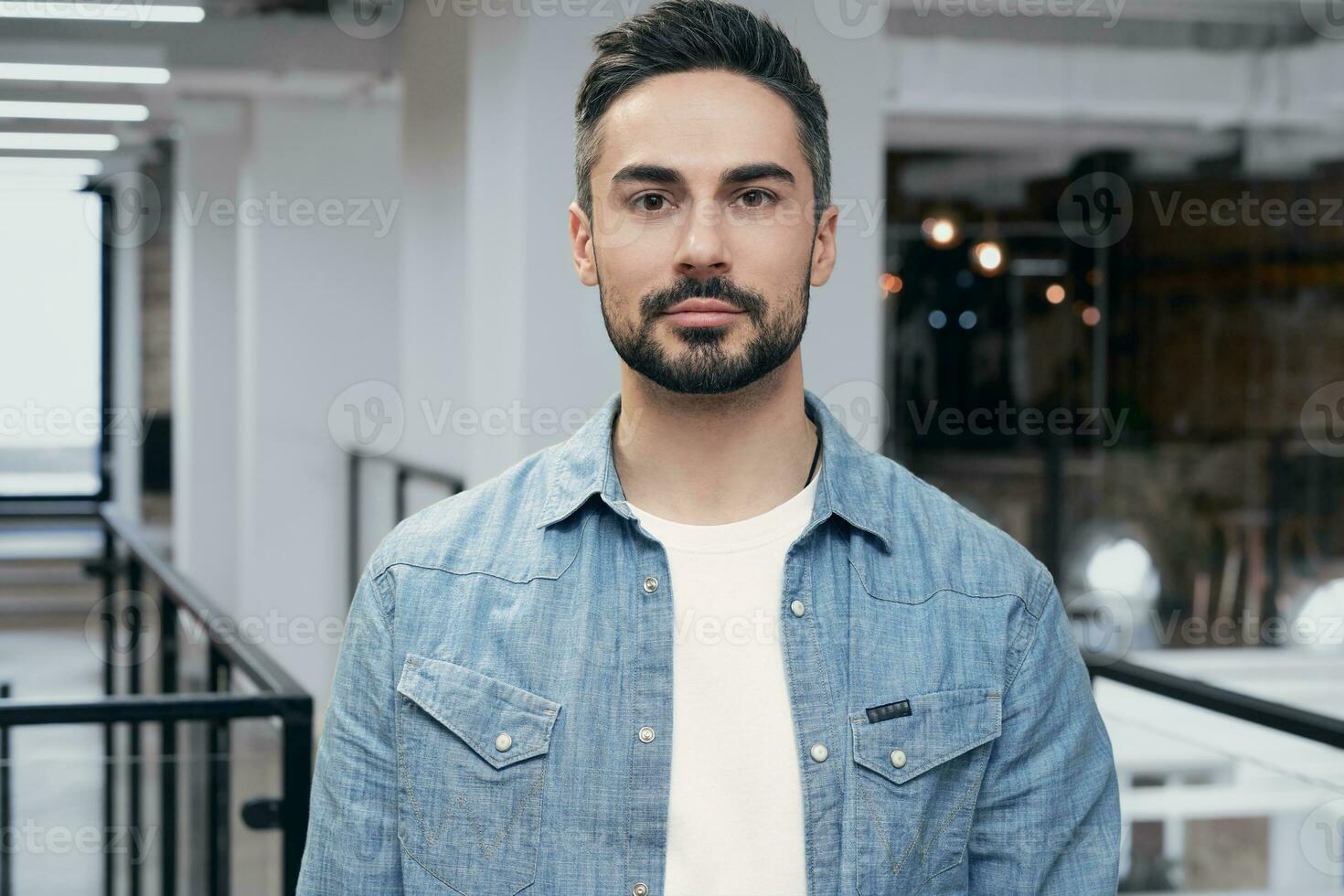 Portrait of young bearded man standing in the office while posing at camera. Modern coworking on the background. Startup, coworking, management concept photo