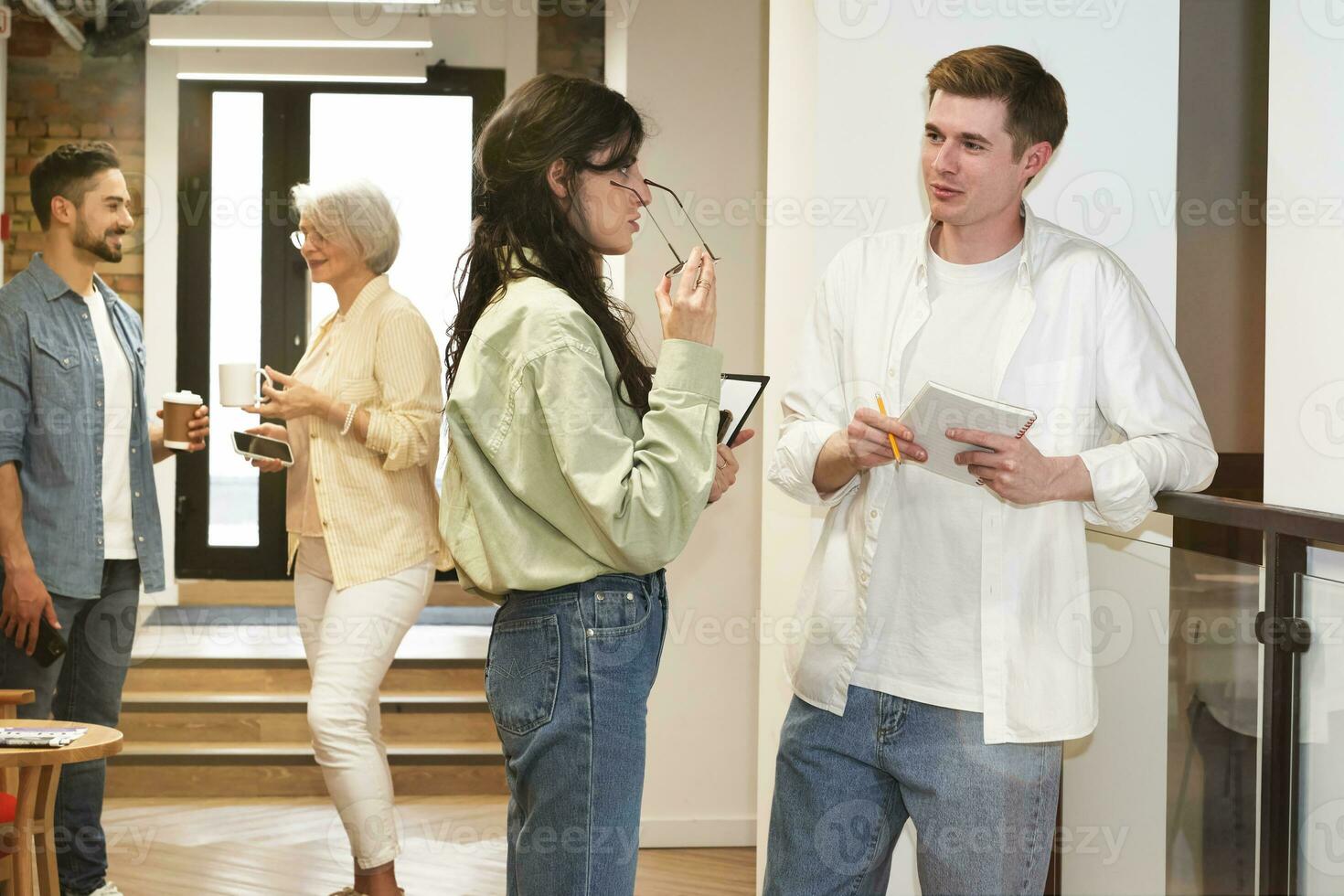 Two colleagues standing in the corridor and discussing job, employees standing with coffee on the background. Startup, coworking, management concept photo