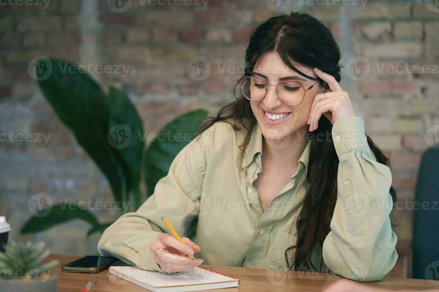 Happy pretty woman sitting at her workplace while writing in notebook in the office. Startup, coworking, management concept photo