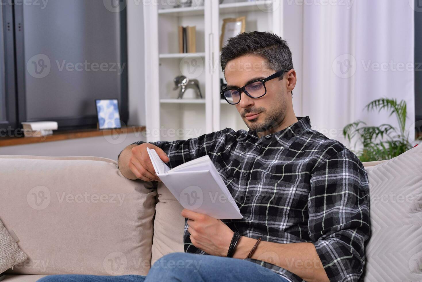 An intellectual young man, donning stylish glasses, immerses himself in a captivating book. With cozy decor, he relishes a tranquil reading session on the sofa in his charming home. photo