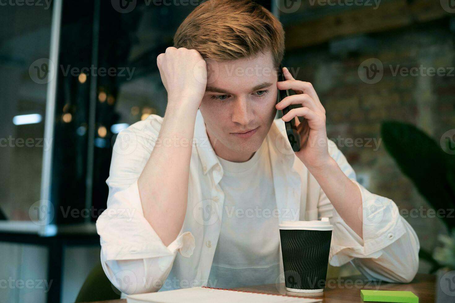 Young man sitting at his workplace in the office while talking on mobile phone, coffee on the table. Startup, coworking, management concept photo