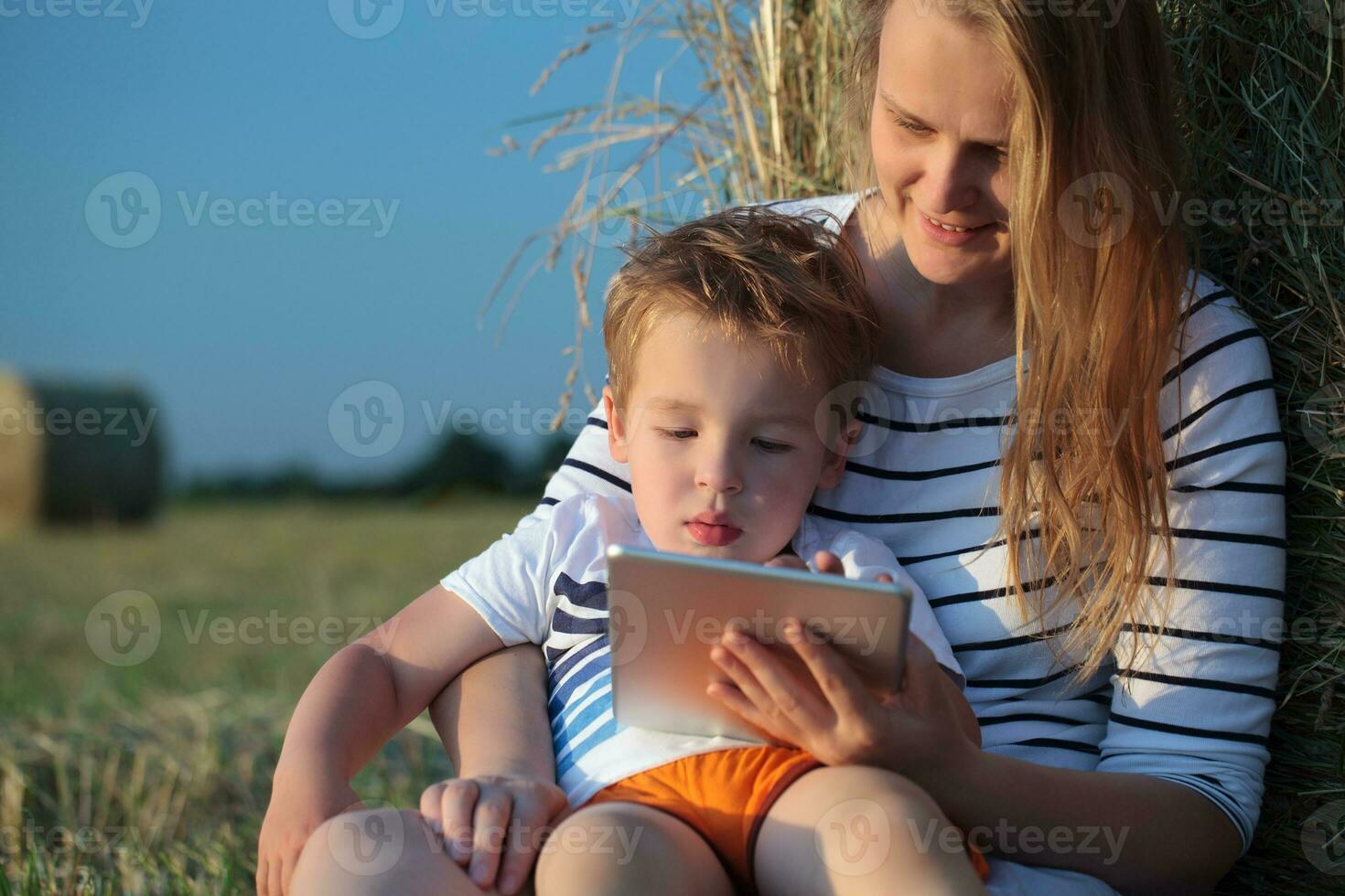 Mother and son with pad sitting by hay roll in the field photo