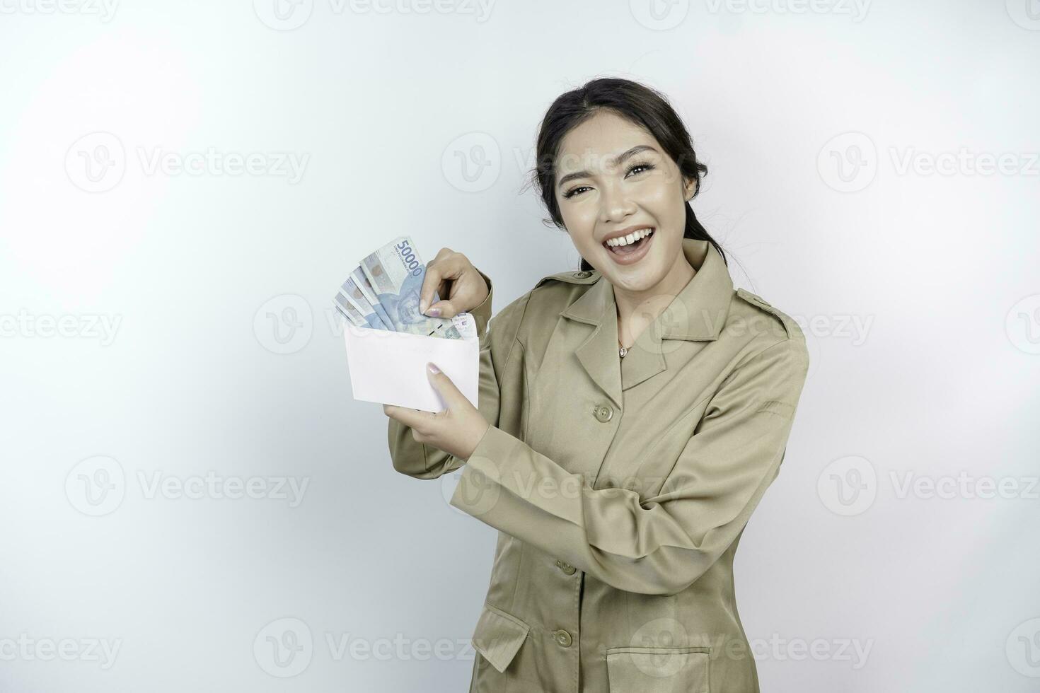 A smiling young government worker woman is wearing khaki uniform holding cash money in Indonesian rupiah isolated by white background photo