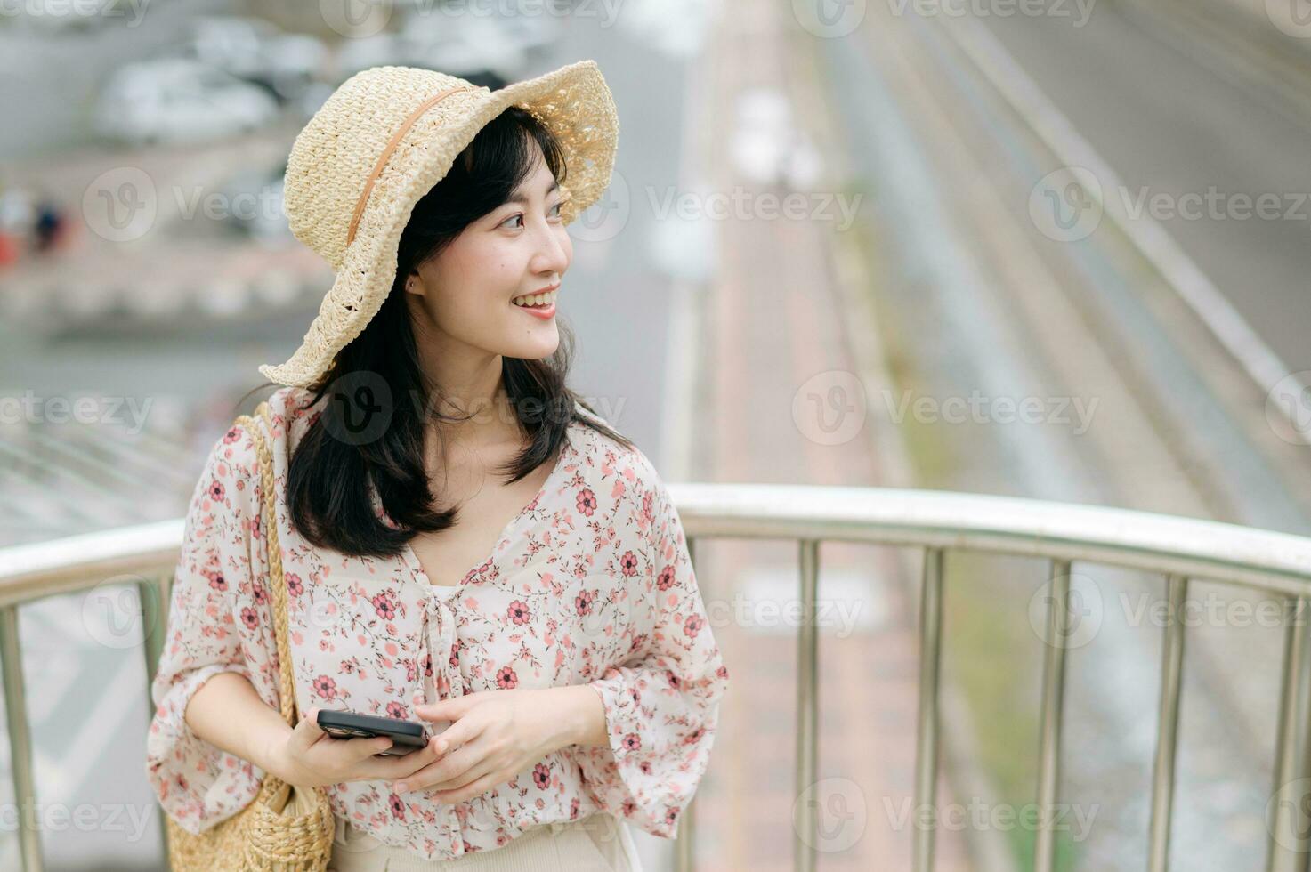 joven asiático mujer viajero con Costura cesta utilizando móvil teléfono y en pie en paso superior con ferrocarril antecedentes. viaje viaje estilo de vida, mundo viaje explorador o Asia verano turismo concepto. foto