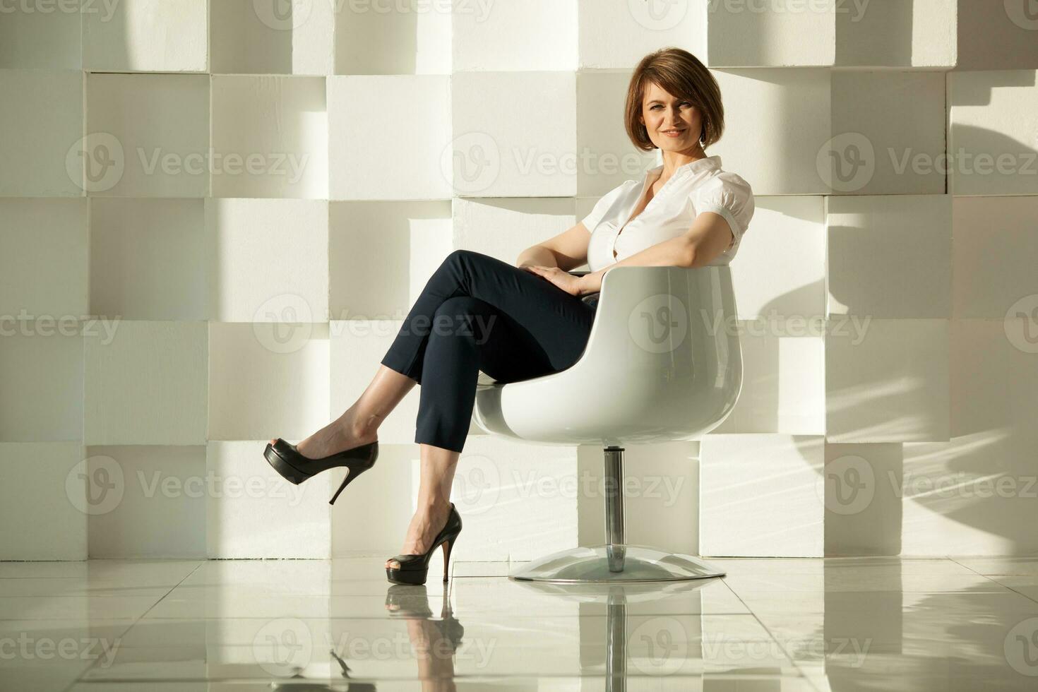 Elegant adult woman sitting in chair against of white modern wall photo