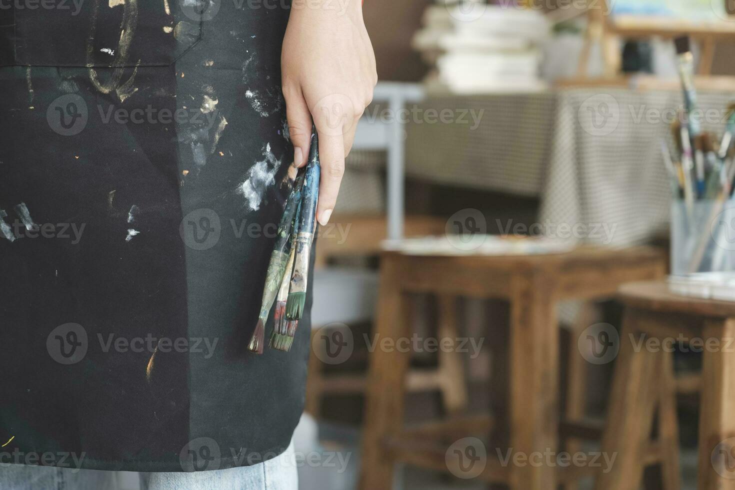 A close-up of the artist hands wearing an apron smeared with paint. Clutching many brushes and paintbrushes. photo