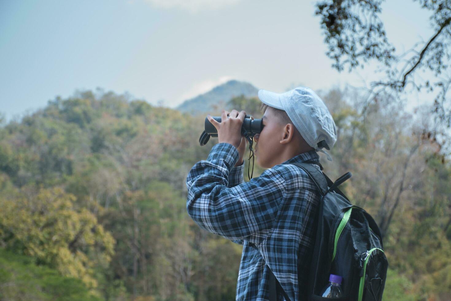 asiático chico en tartán camisa y gorra haciendo el verano actividad a local nacional parque por acecho aves, pez, insectos, animales, árboles, flor por utilizando mapa y prismáticos, suave y selectivo enfocar. foto