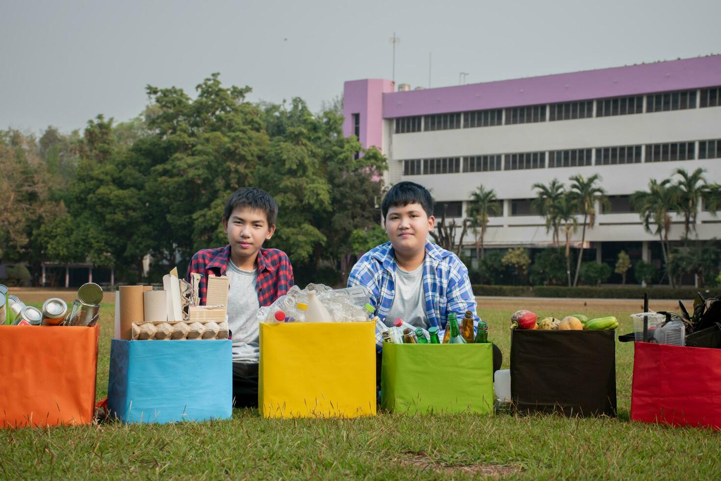 Asian boy in plaid shirt sitting on playground of school with various boxes of separated garbabes or trashes inside, concept for eco friends and global warming campaign, soft and selective focus. photo