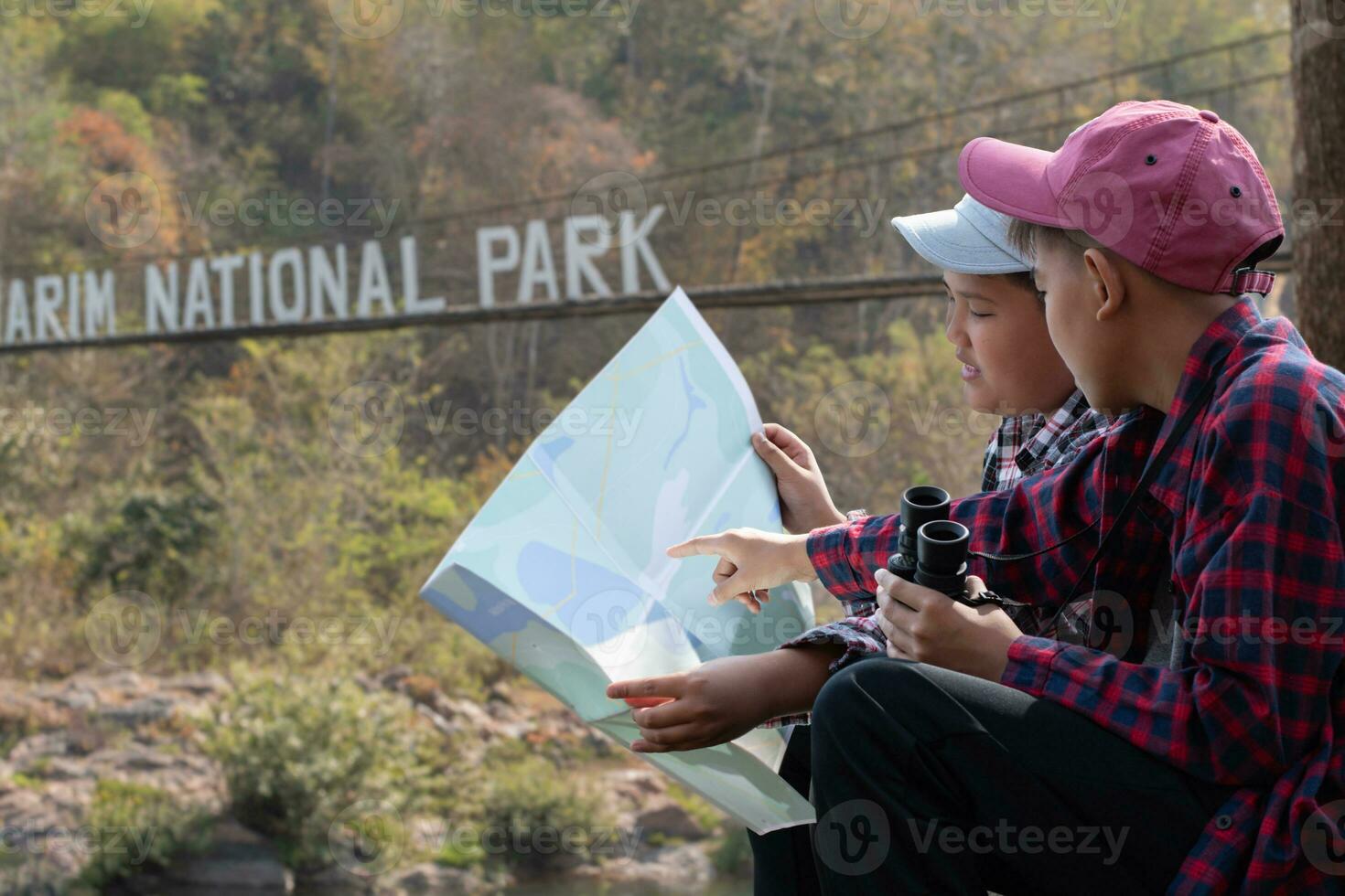 asian boys holds binoculars and local national park map, resting and reading information in map near sling bridge of local national park during nature and summer camp of them, teens' activity concept. photo