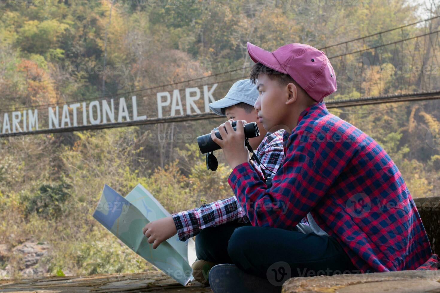 asian boys holds binoculars and local national park map, resting and reading information in map near sling bridge of local national park during nature and summer camp of them, teens' activity concept. photo