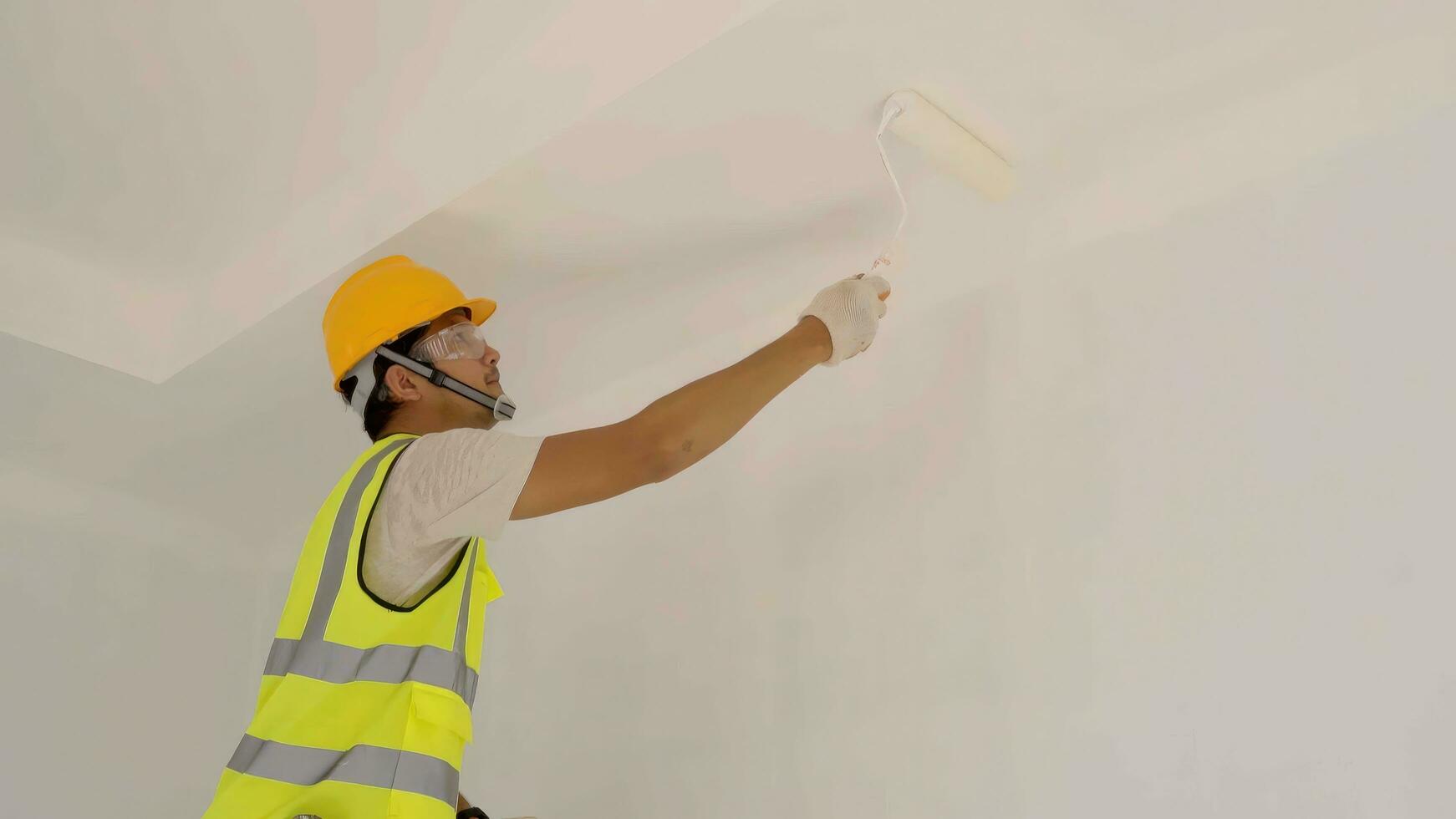 asiático construcción trabajador usos rodillo a pintar el techo a el construcción sitio . foto