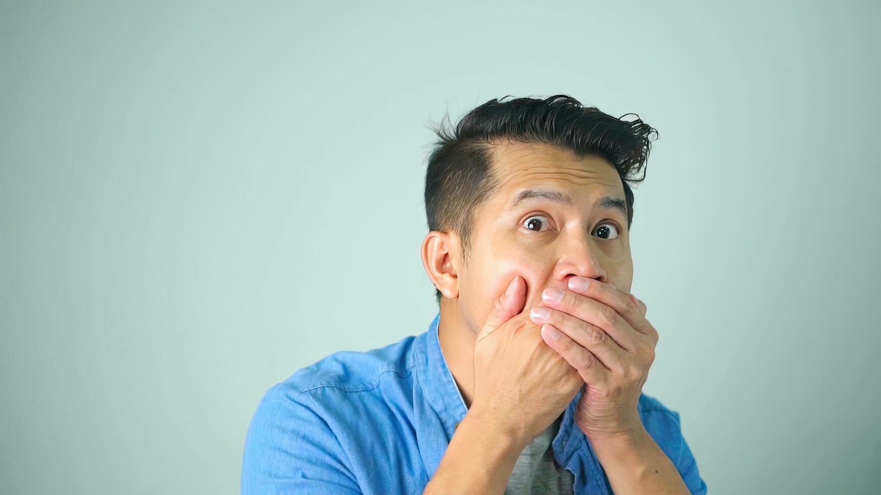 Asian man confused shocked close mouth with hands from unexpected bad news failure crime covers lips oops say terrible secret keep silence. photo