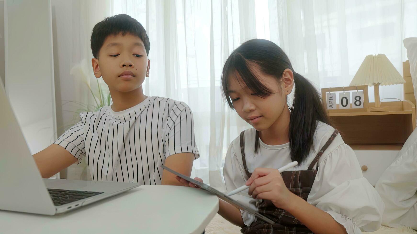 Boy using laptop looking at girl using pen write tablet in bedroom. photo