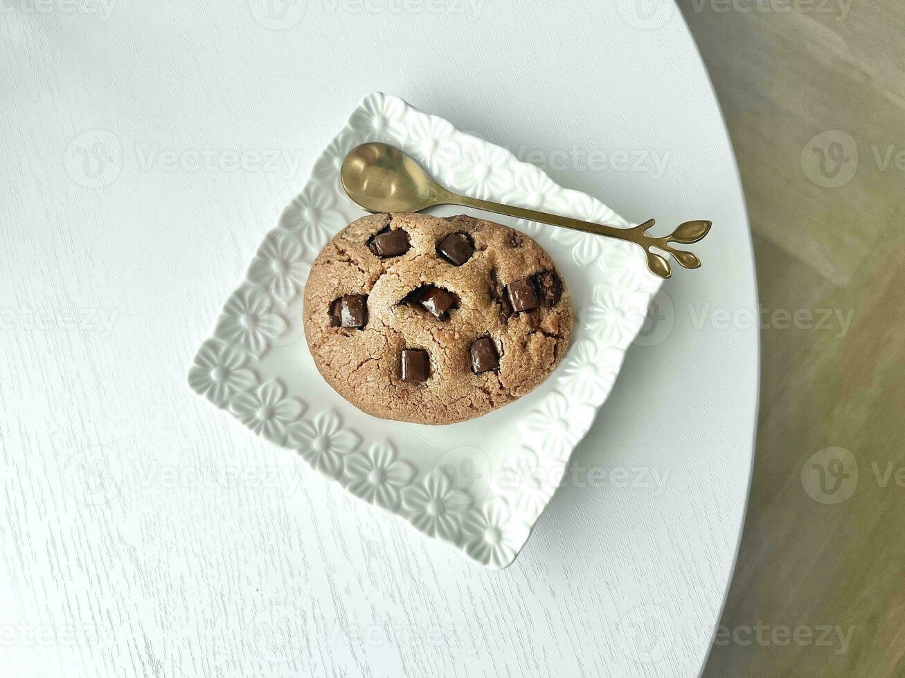 Chocolate chip cookie on a white plate with a golden spoon on white wooden table, top view photo