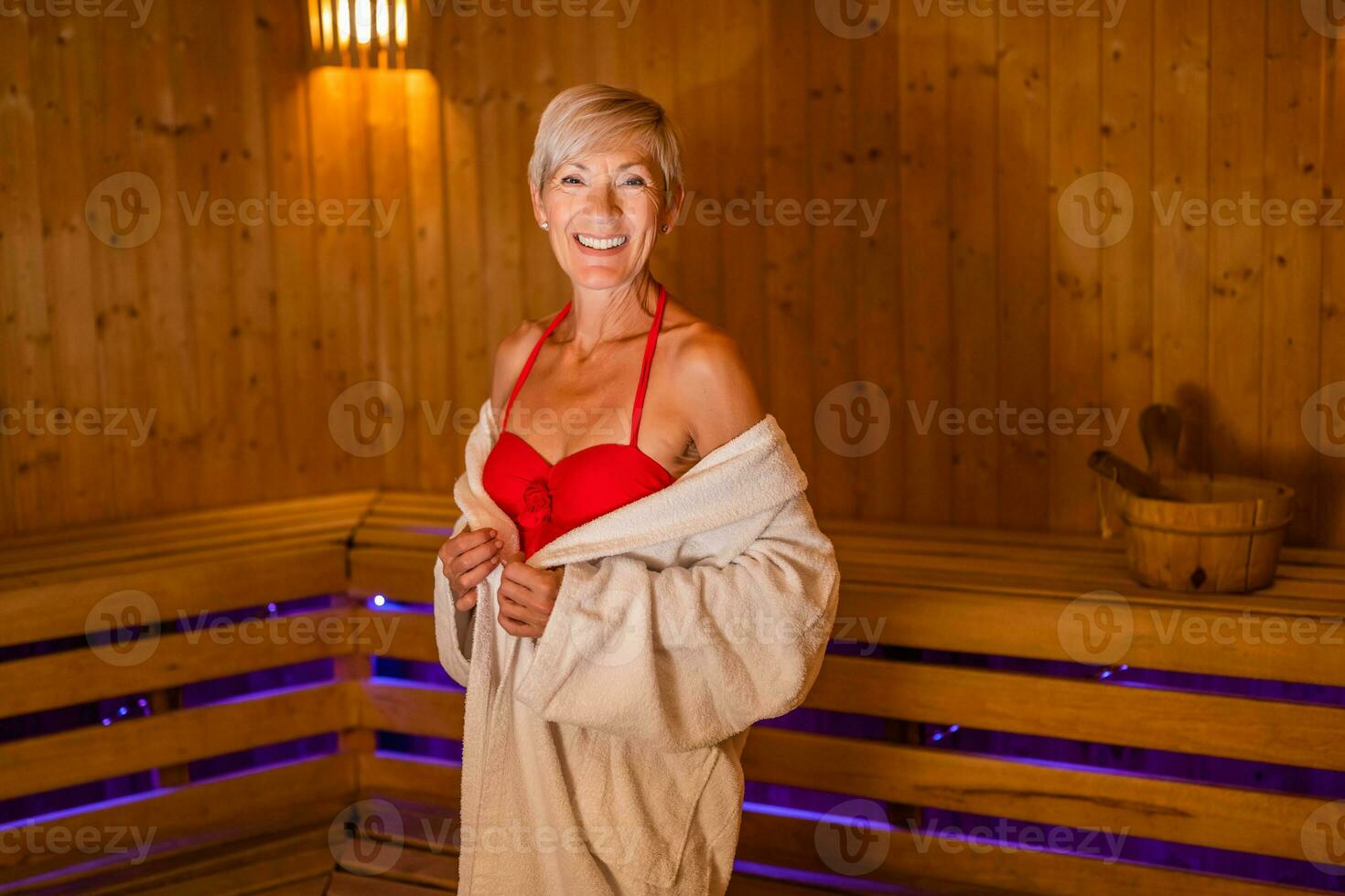 Mature woman is ready for relaxing in sauna. She is standing in sauna and looking at camera. Spa concept. photo