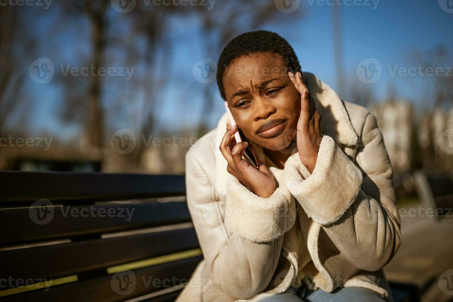 al aire libre retrato de preocupado afroamericano mujer en soleado día. ella es sentado en banco en el calle y hablando en teléfono. foto