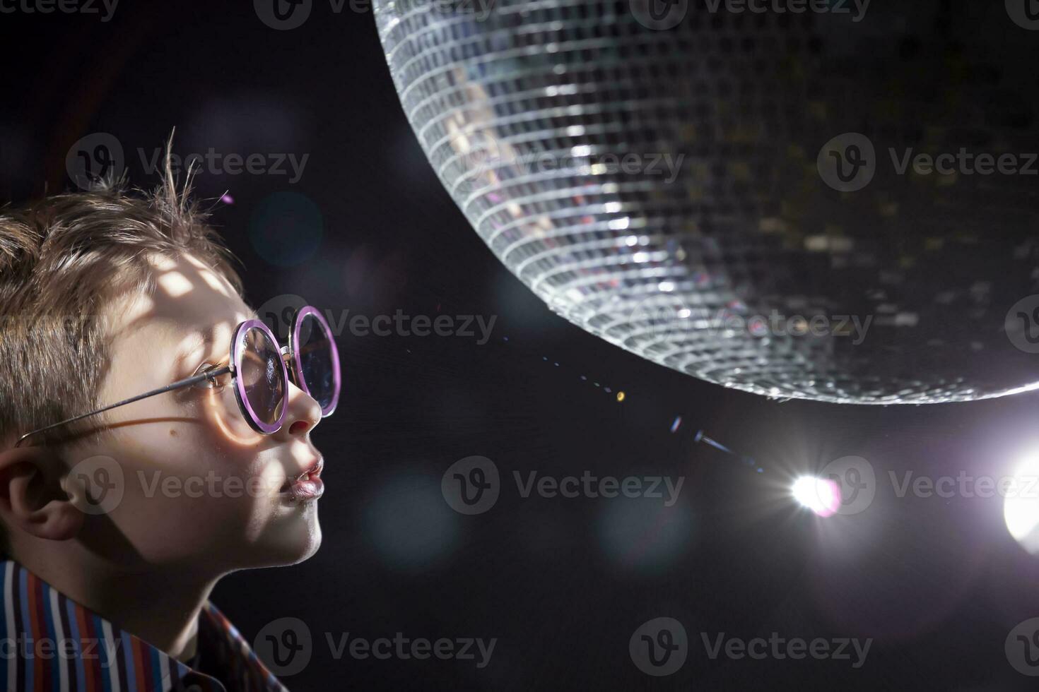 A young boy with glasses looks at a spinning disco ball. Children's disco, party. photo