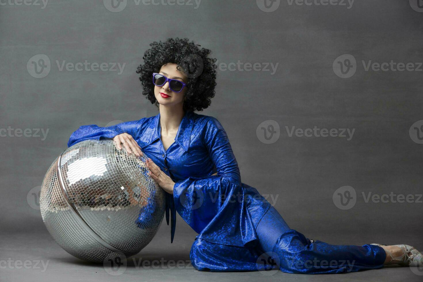 Spectacular Woman in a sparkly flared suit and afro wig poses with a disco ball. Disco style from the seventies or eighties. photo