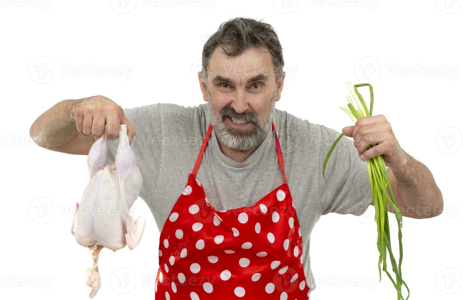 un mayor barbado masculino cocinar en un rojo lunares delantal sostiene un crudo pollo y un manojo de cebollas en un blanco antecedentes. cocinar con pollo. foto