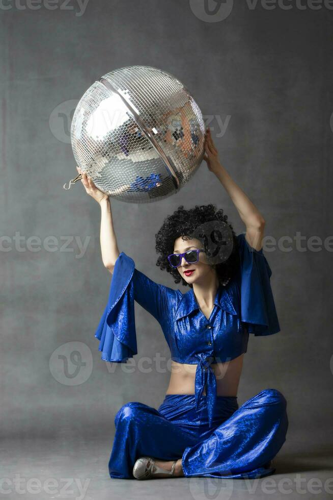 A woman in a seventies flared suit with a disco ball in an afro wig on a gray background. photo
