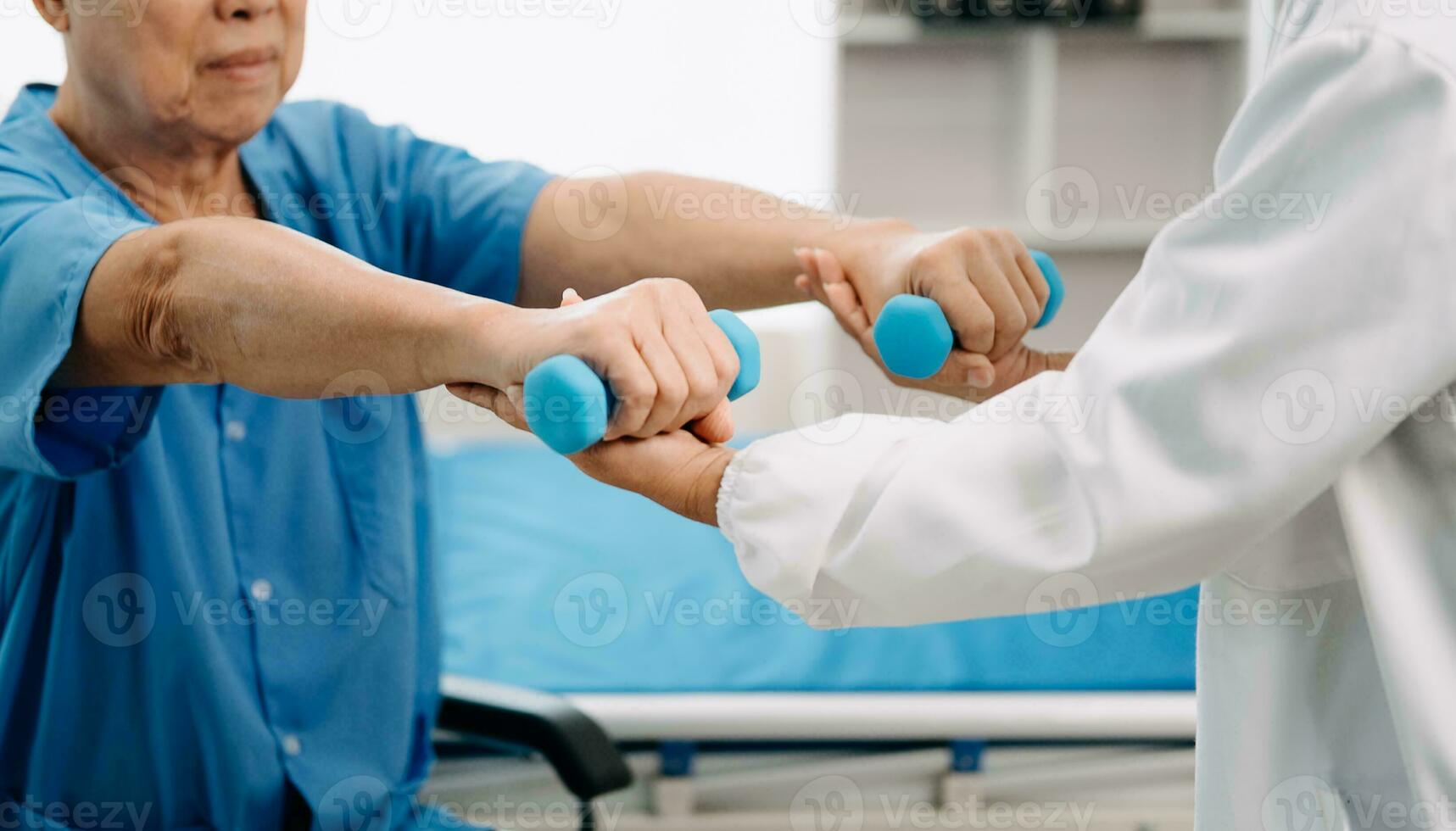 Asian physiotherapist helping elderly man patient stretching arm during exercise correct with dumbbell in hand during training hand in bed in clinic photo