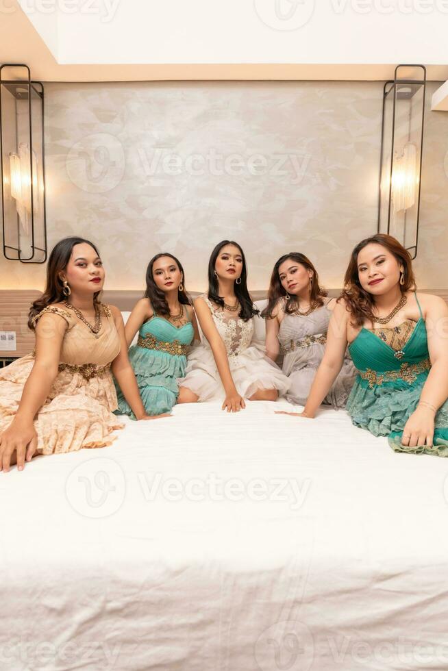 un grupo de asiático mujer en hermosa vestidos sentado juntos en un blanco cama mientras quedarse foto