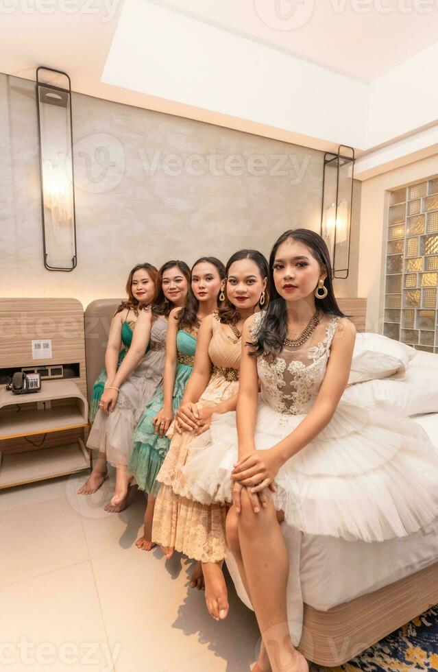 a group of Asian women sitting together on a white bed while wearing dresses and makeup during a party at a friend's house photo