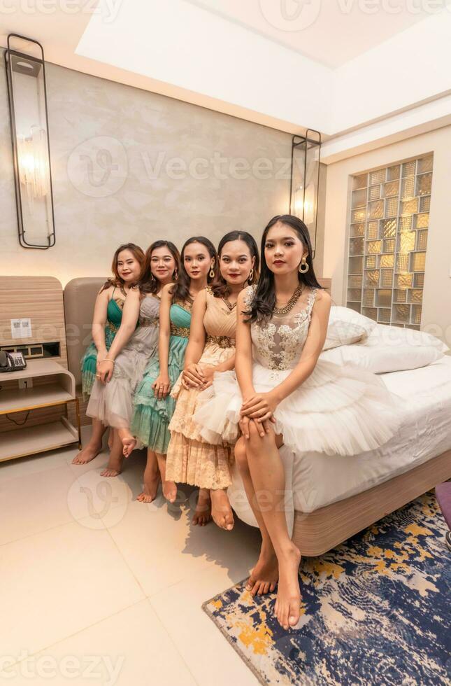 un grupo de asiático mujer sentado juntos en un blanco cama mientras vistiendo vestidos y maquillaje durante un fiesta a un amigos casa foto