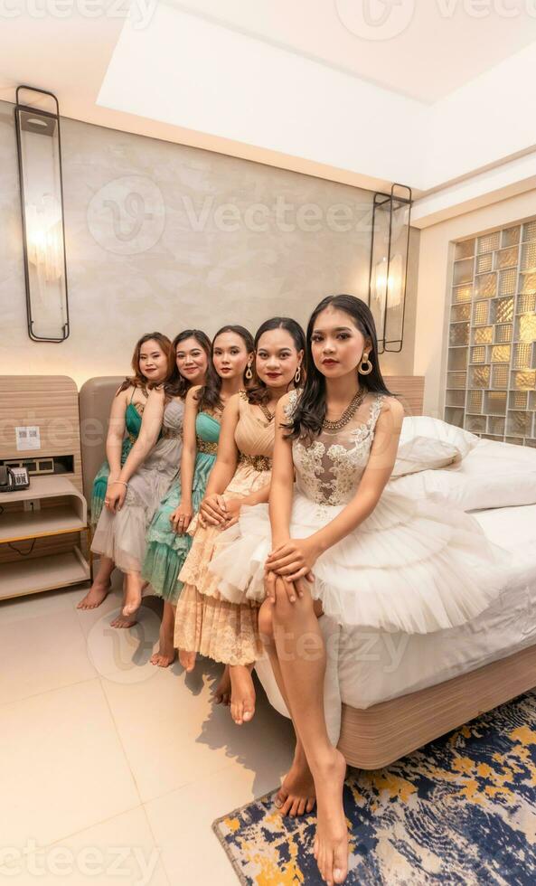 a group of Asian women sitting together on a white bed while wearing dresses and makeup during a party at a friend's house photo