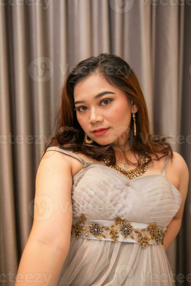 un asiático mujer es posando muy hermosamente mientras vistiendo un oro vestir y pendientes a un Boda foto