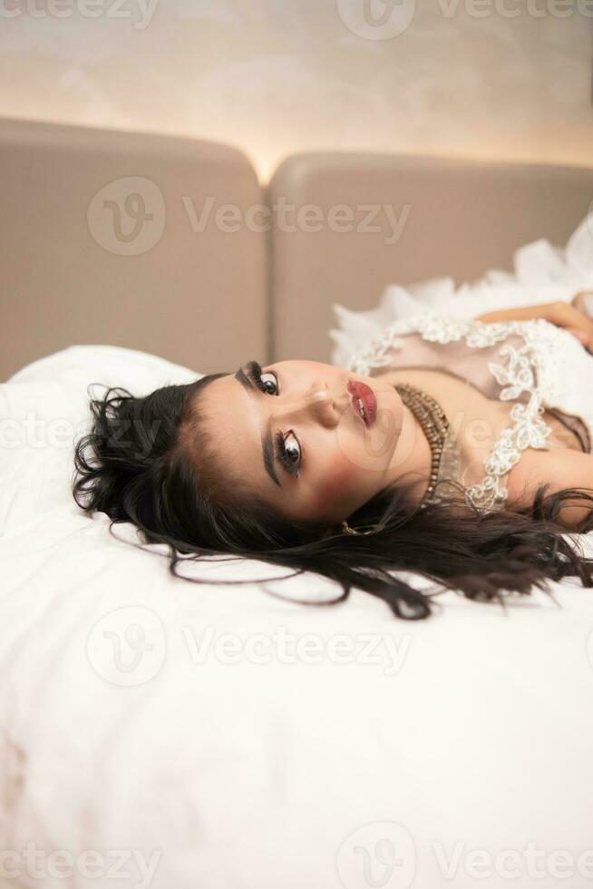 un asiático mujer con negro pelo mordiendo su dedos con un córneo expresión mientras dormido en un blanco cama foto