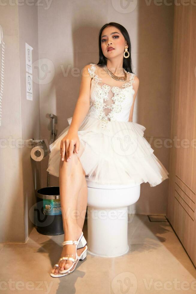 un asiático mujer en un blanco vestir y hermosa maquillaje es sentado esmeradamente en un blanco baño en un lujoso hotel habitación foto