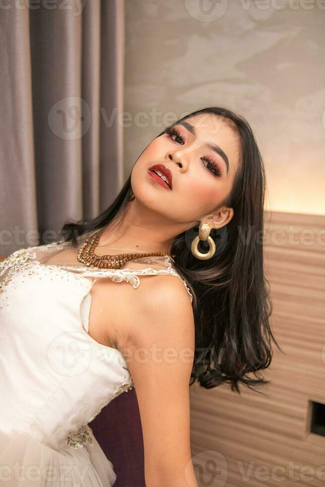 an Asian woman in a sexy white dress is posing on a purple sofa in a hotel photo