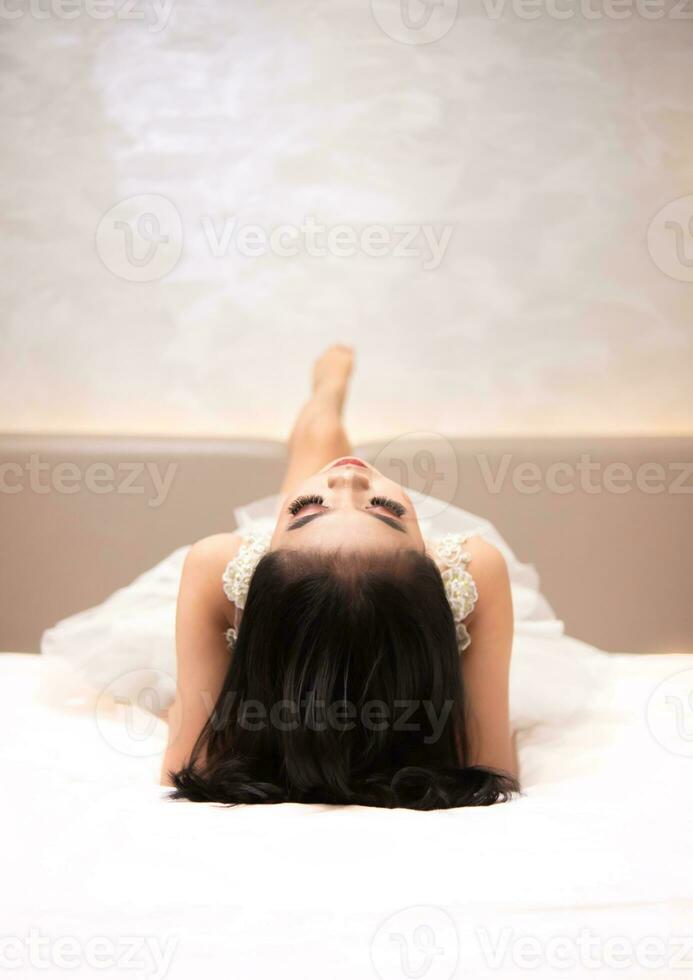 an Asian woman in a white dress is lying on a white bed with her black hair loose in a hotel photo