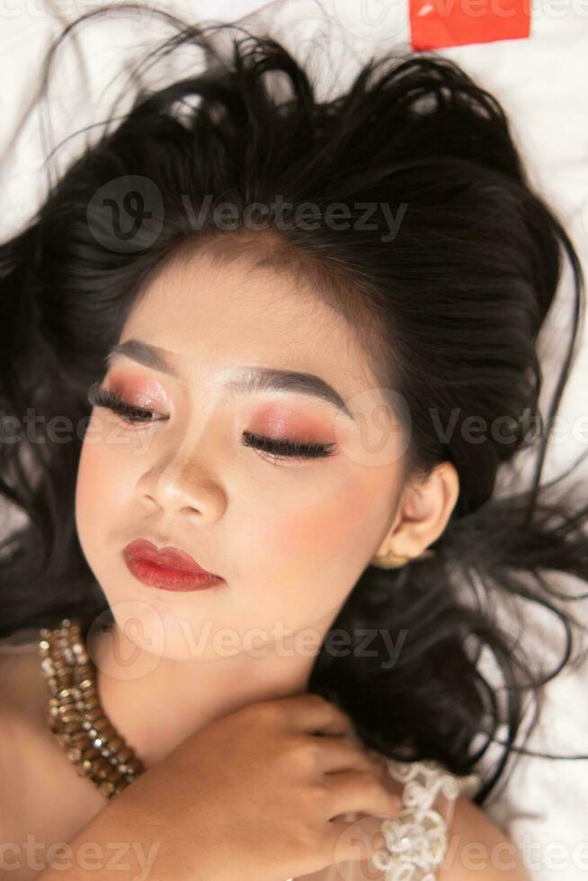 an Asian woman with beautiful makeup sleeping on a white bed while wearing a dress photo