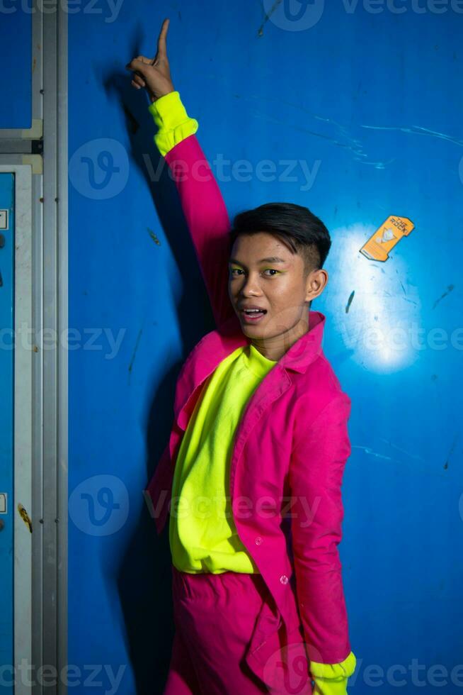 a gay Asian man posing very funny and humorous while wearing a pink suit in front of a blue background photo