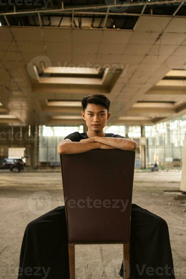 un asiático hombre posando en un marrón cuero silla mientras vistiendo negro pantalones en un antiguo edificio foto