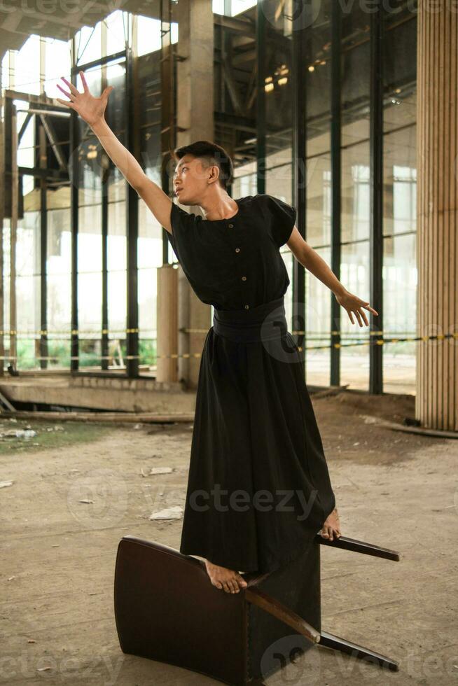 an Asian man is standing on a wooden chair while wearing a black dress in an old building photo