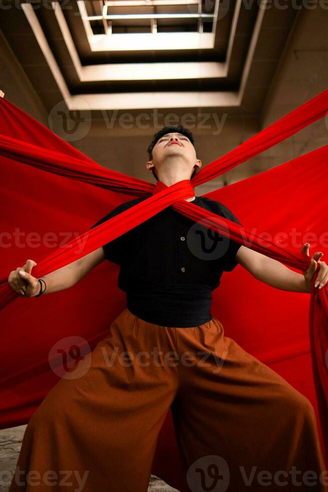 an Asian man stands under the light as his body is bound by a red cloth photo