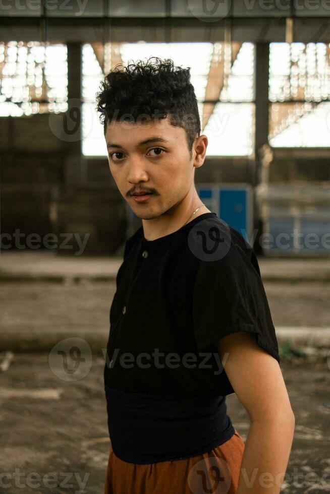 an Asian man in a black shirt and brown pants standing proudly in front of an old building photo