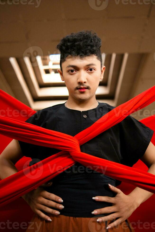 an Asian man stands under the light as his body is bound by a red cloth photo