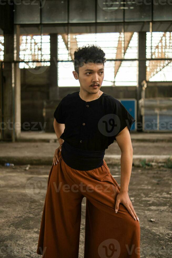 an Asian man in a black shirt and brown pants standing proudly in front of an old building photo