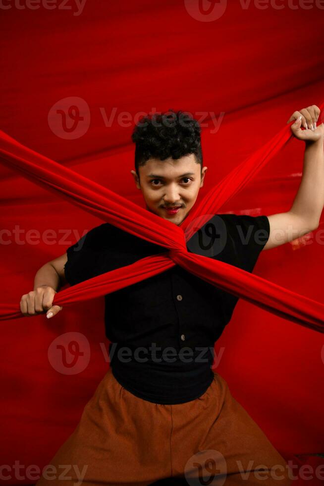 un asiático hombre soportes en frente de un rojo en forma de x paño y sostiene eso con ambos manos foto