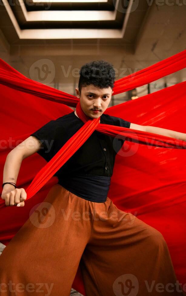 an Asian man in a black shirt is strangled with a red cloth while standing in an old building against a red background photo